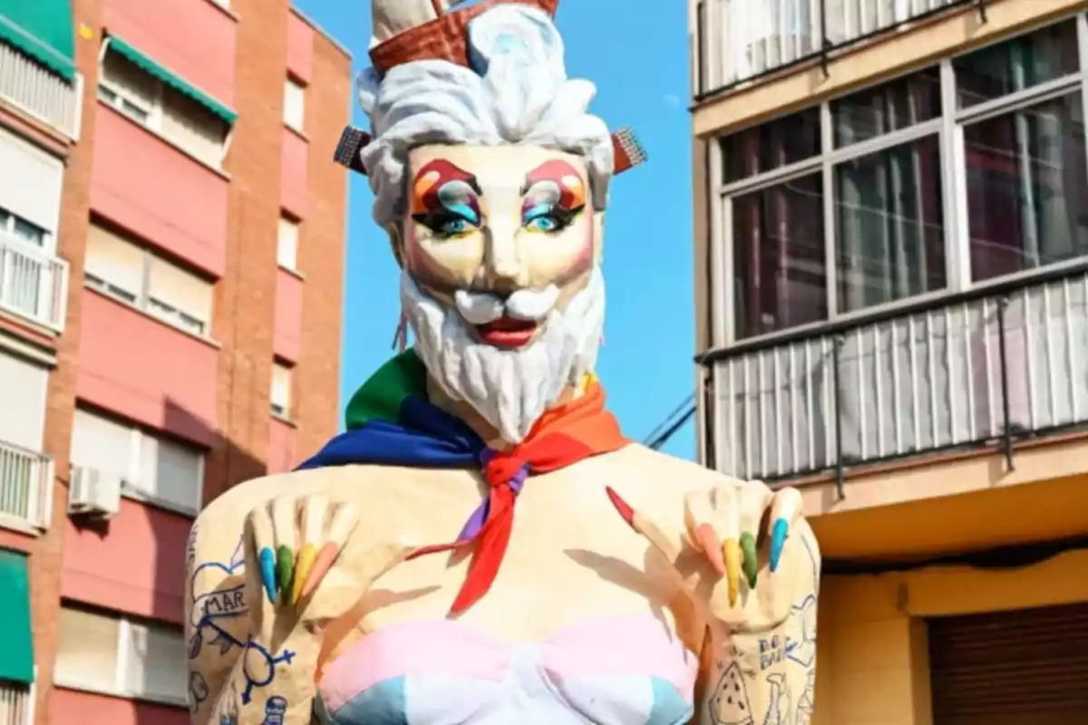Figura de cartón con barba blanca, maquillaje colorido, uñas pintadas de colores y pañuelo arcoíris en el cuello, ubicada en una calle entre edificios.