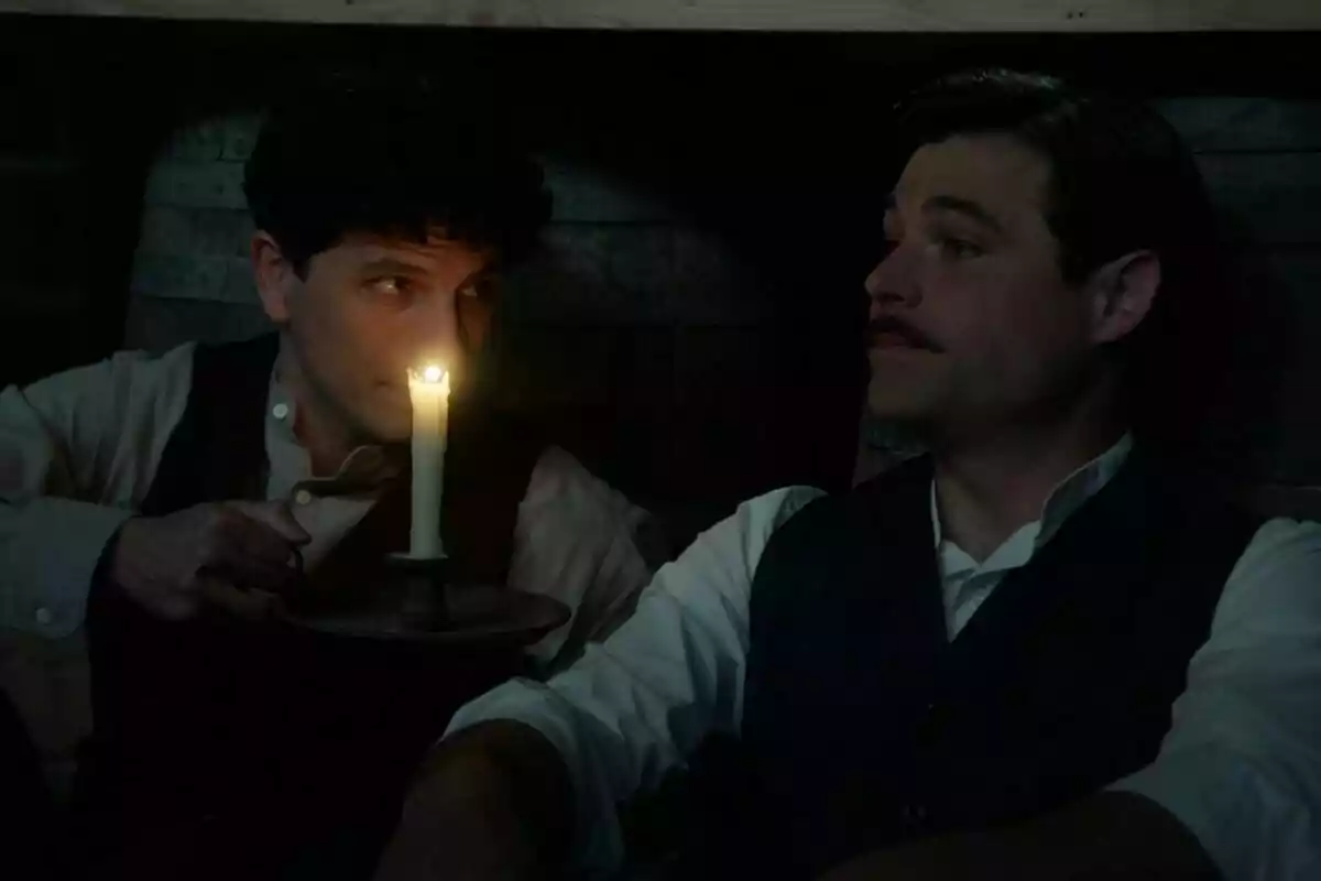 Dos hombres sentados en un lugar oscuro, Álvaro Quintana sujetando una vela mientras mira a Arturo Sancho en La Promesa Dos hombres sentados en un lugar oscuro, Álvaro Quintana sujetando una vela mientras mira a Arturo Sancho en La Promesa