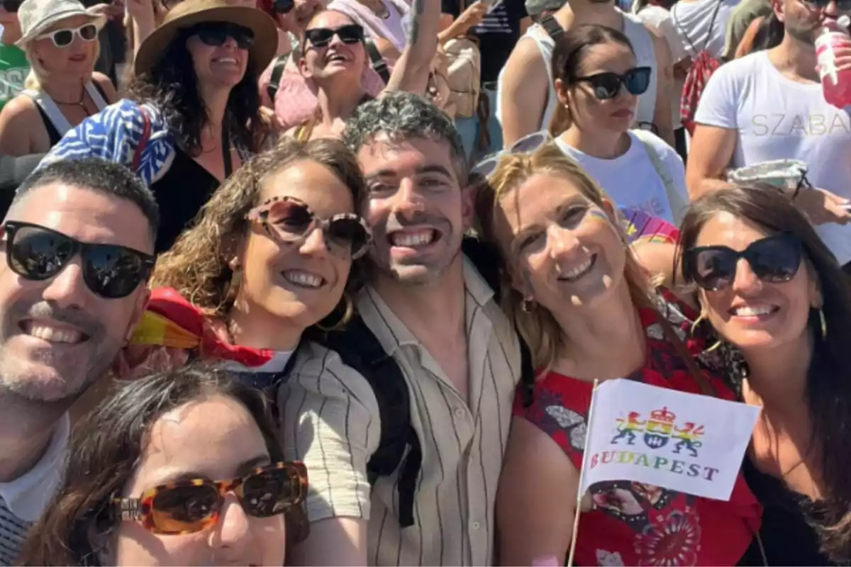 Grupo de personas sonrientes con gafas de sol disfrutando de un evento al aire libre, una de ellas sostiene una bandera que dice Budapest