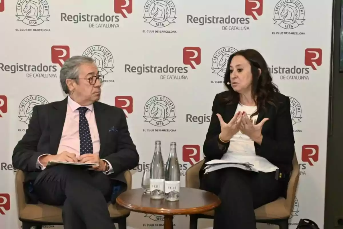 Dos personas sentadas conversan en un evento organizado por Registradores de Catalunya y Círculo Ecuestre, con una mesa pequeña y botellas de agua entre ellas. Dos personas sentadas conversan en un evento organizado por Registradores de Catalunya y Círculo Ecuestre, con una mesa pequeña y botellas de agua entre ellas.