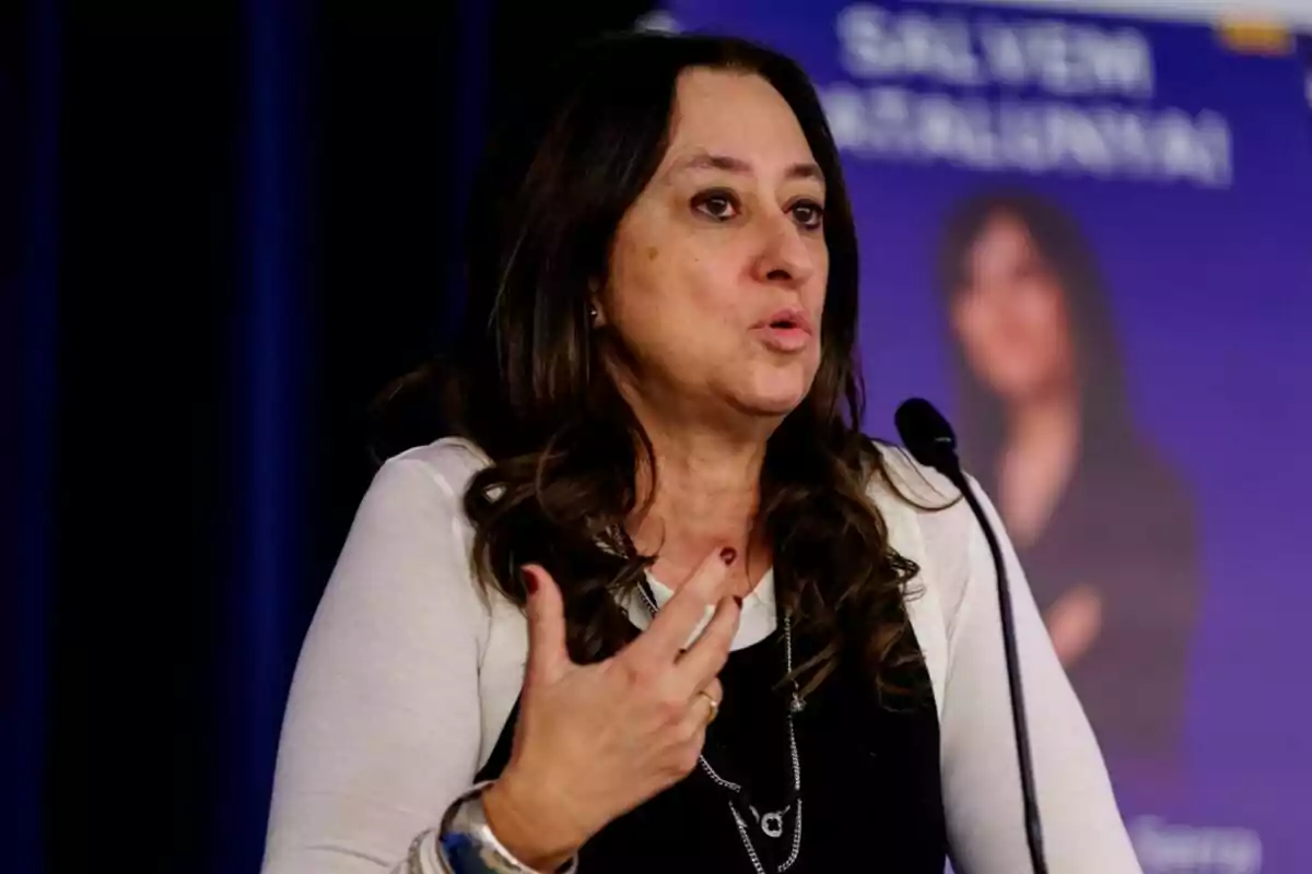 Mujer de cabello largo y oscuro hablando frente a un micrófono con un fondo azul y un cartel desenfocado detrás de ella
