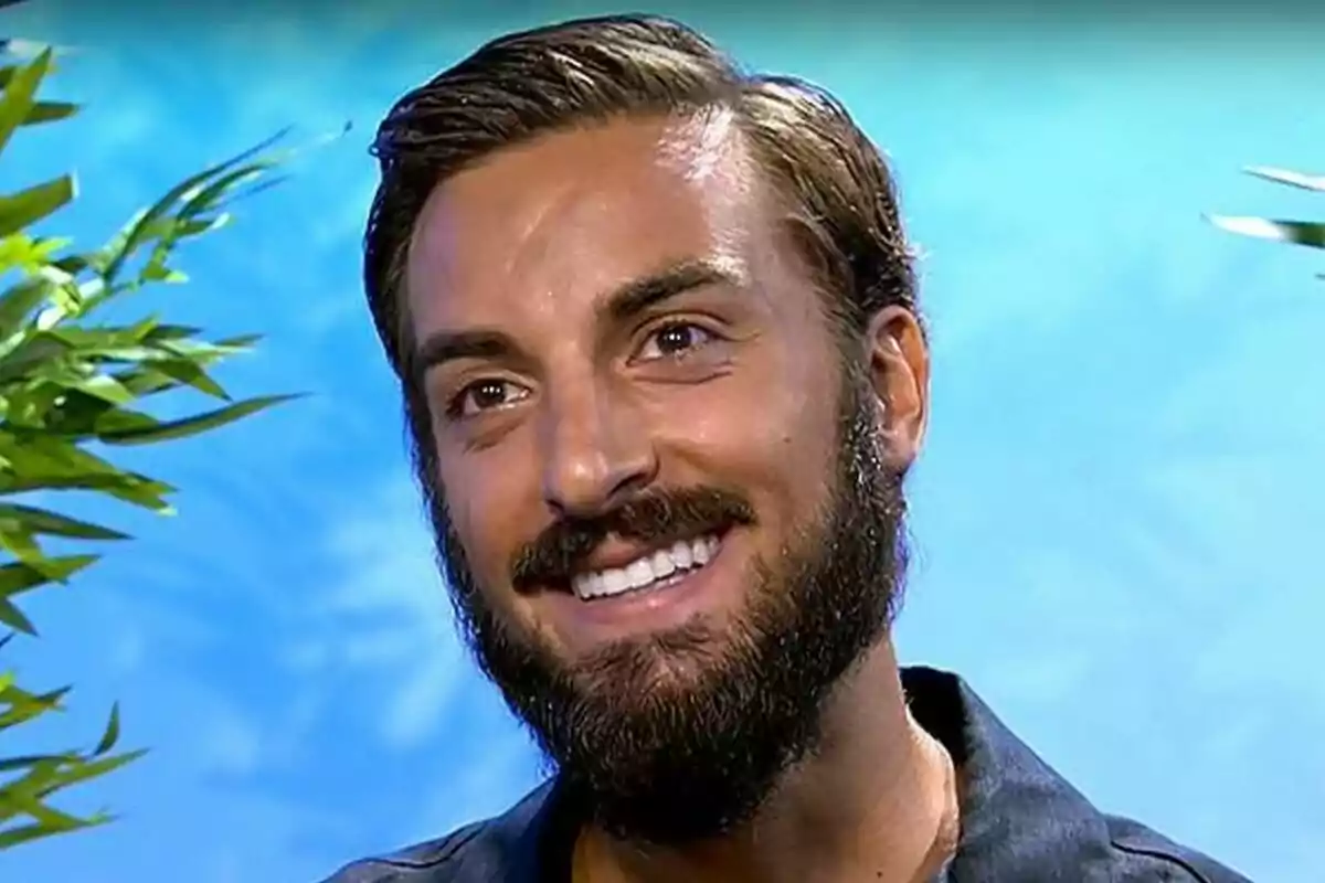 Borja González joven con barba y cabello corto sonríe frente a un fondo azul con plantas a los lados en Supervivientse