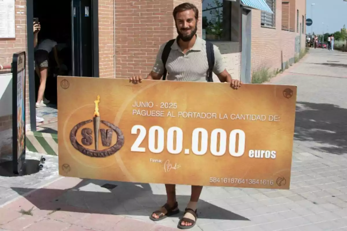 Borja González sonriente sostiene un cheque gigante de 200,000 euros en la calle frente a un edificio de ladrillo Borja González sonriente sostiene un cheque gigante de 200,000 euros en la calle frente a un edificio de ladrillo