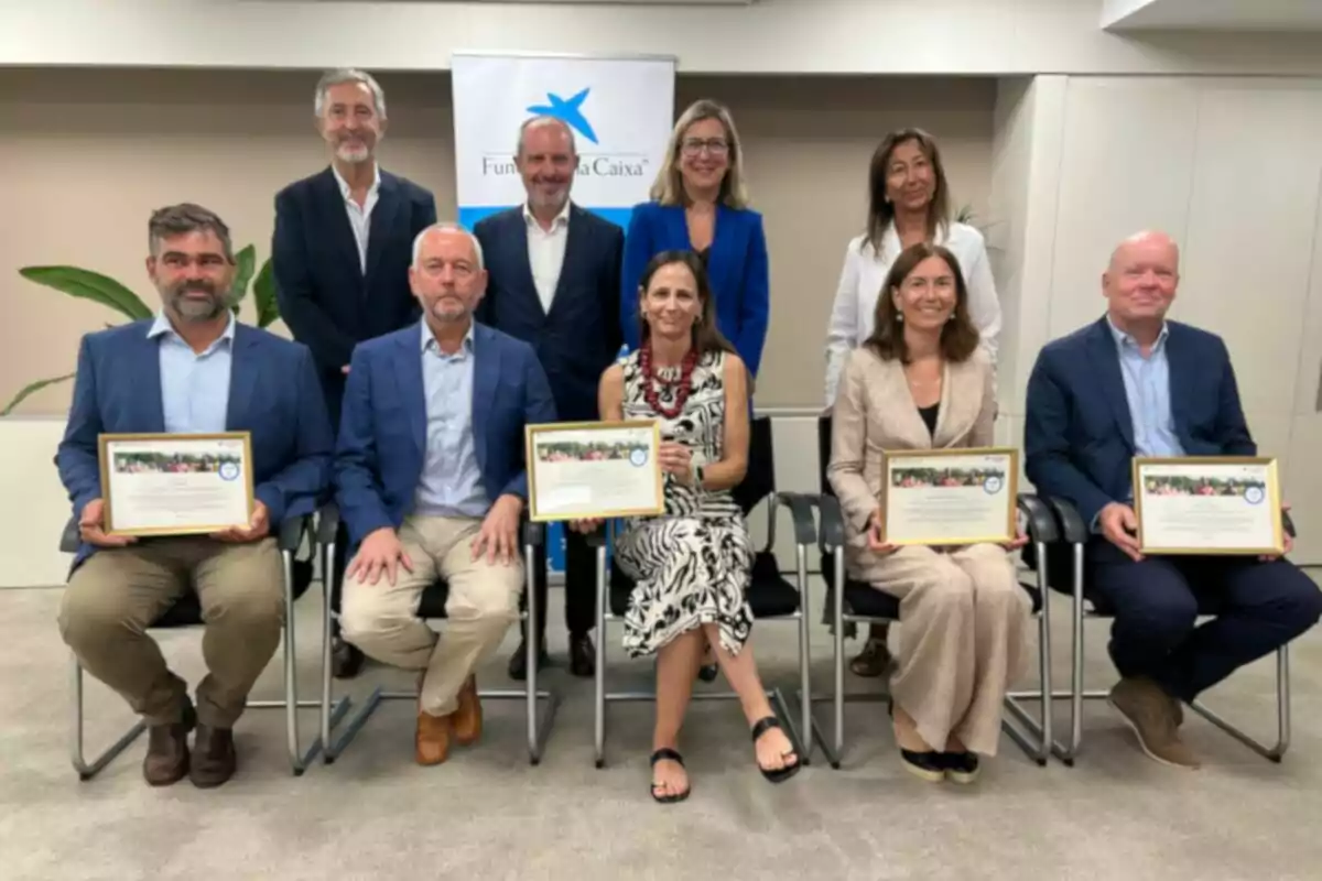 Grupo de personas posando en una sala de oficina, algunas sentadas y otras de pie, varias sostienen diplomas enmarcados y al fondo hay un cartel de Fundación La Caixa Grupo de personas posando en una sala de oficina, algunas sentadas y otras de pie, varias sostienen diplomas enmarcados y al fondo hay un cartel de Fundación La Caixa