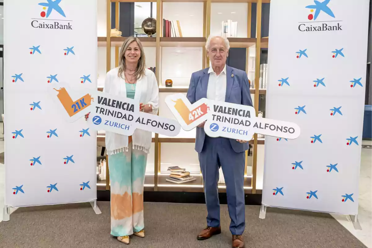 Dos personas posan sonrientes en un evento de CaixaBank sosteniendo carteles promocionales de las carreras 21K y 42K Valencia Trinidad Alfonso Zurich, con un fondo de estantería y paneles con el logo de la entidad bancaria. Dos personas posan sonrientes en un evento de CaixaBank sosteniendo carteles promocionales de las carreras 21K y 42K Valencia Trinidad Alfonso Zurich, con un fondo de estantería y paneles con el logo de la entidad bancaria.