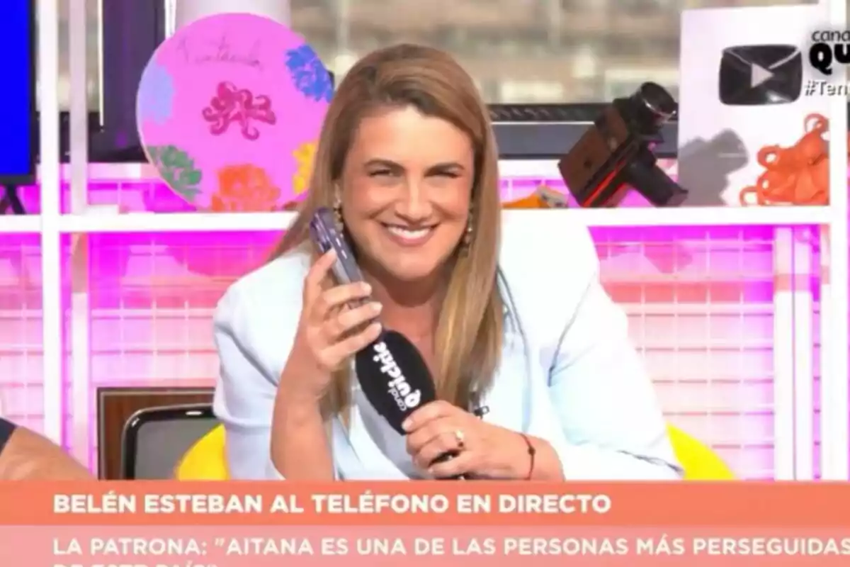Carlota Corredera sonriente en 'Tentáculos' sosteniendo un teléfono móvil y un micrófono mientras está sentada en un set colorido con objetos decorativos al fondo Carlota Corredera sonriente en 'Tentáculos' sosteniendo un teléfono móvil y un micrófono mientras está sentada en un set colorido con objetos decorativos al fondo