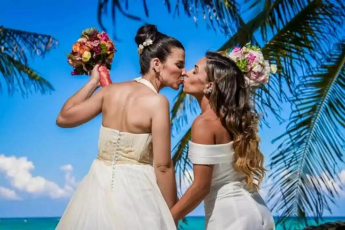 Dos mujeres vestidas de novia se besan en la playa bajo palmeras y un cielo azul, ambas sostienen ramos de flores coloridas en Casados a primera vista. Dos mujeres vestidas de novia se besan en la playa bajo palmeras y un cielo azul, ambas sostienen ramos de flores coloridas en Casados a primera vista.