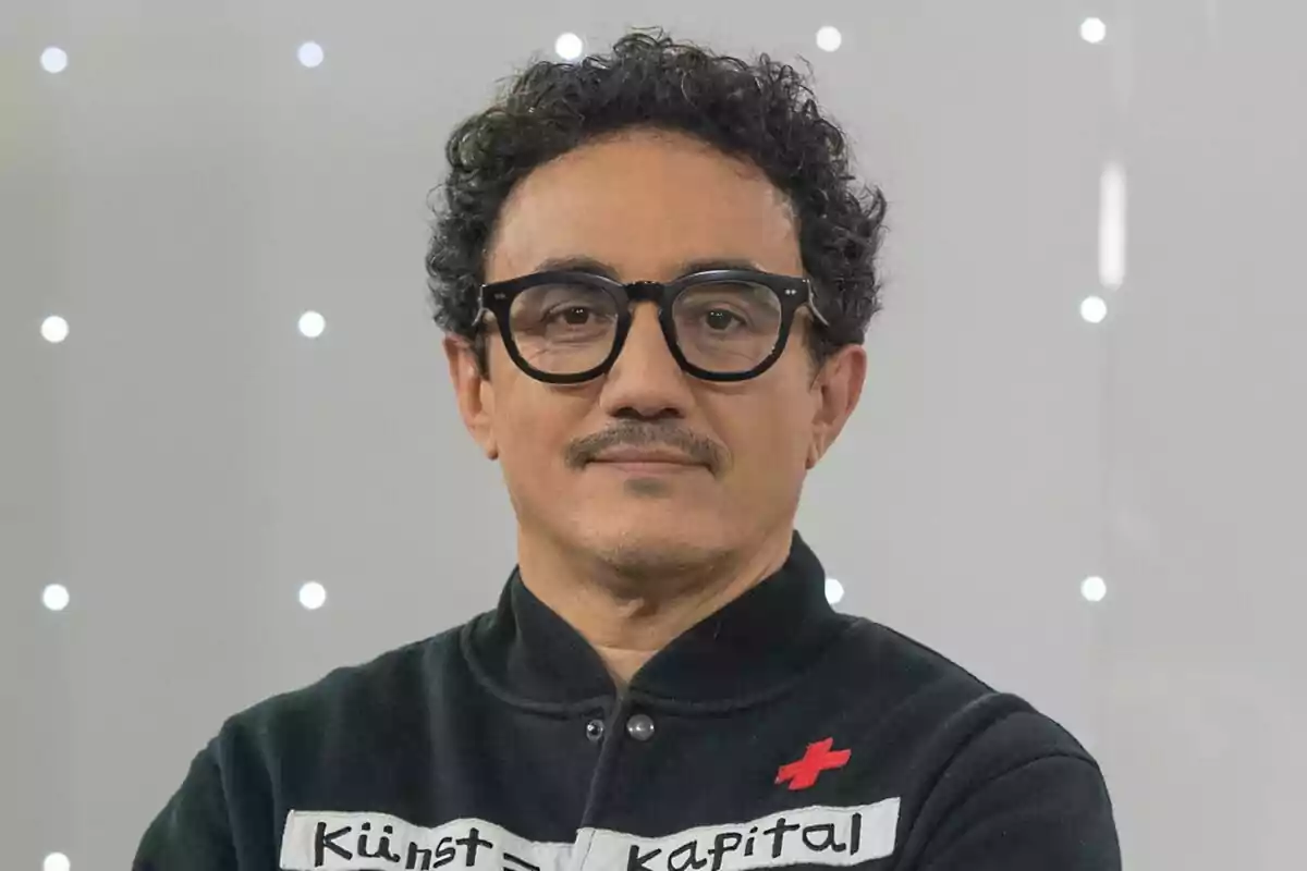 César Vallejo de cabello rizado y gafas negras con expresión seria, usando un suéter oscuro con letras y fondo de luces blancas desenfocadas