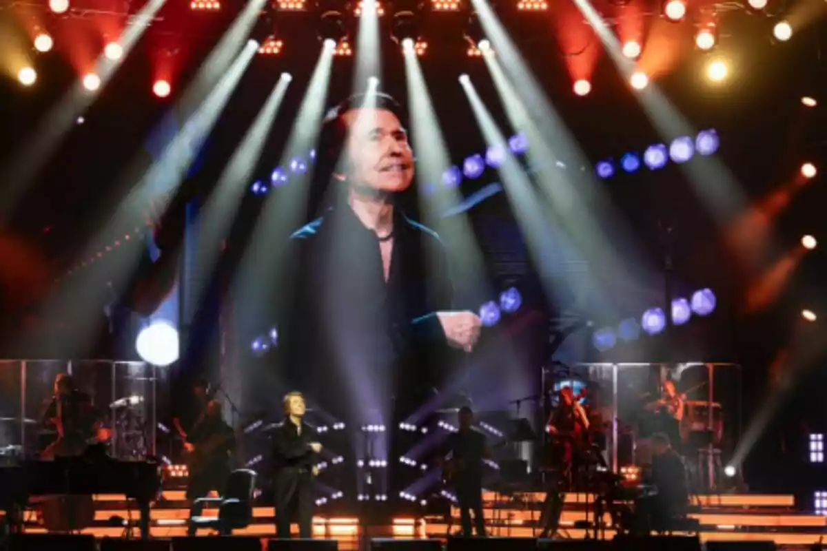 Raphael se presenta en un escenario iluminado con luces de colores y una gran pantalla al fondo muestra su imagen mientras la banda toca detrás de él. Raphael se presenta en un escenario iluminado con luces de colores y una gran pantalla al fondo muestra su imagen mientras la banda toca detrás de él.