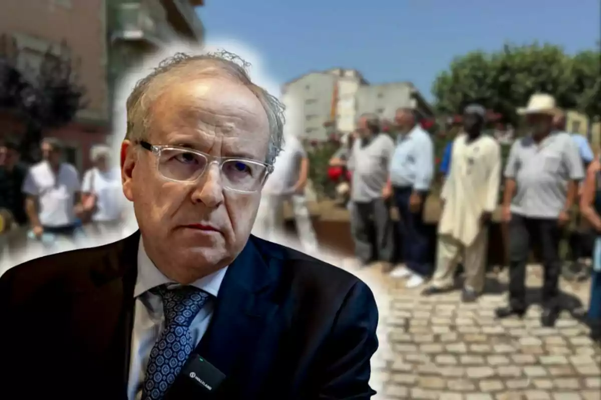 Hombre de traje y gafas en primer plano con expresión seria, al fondo un grupo de personas desenfocadas en una calle urbana
