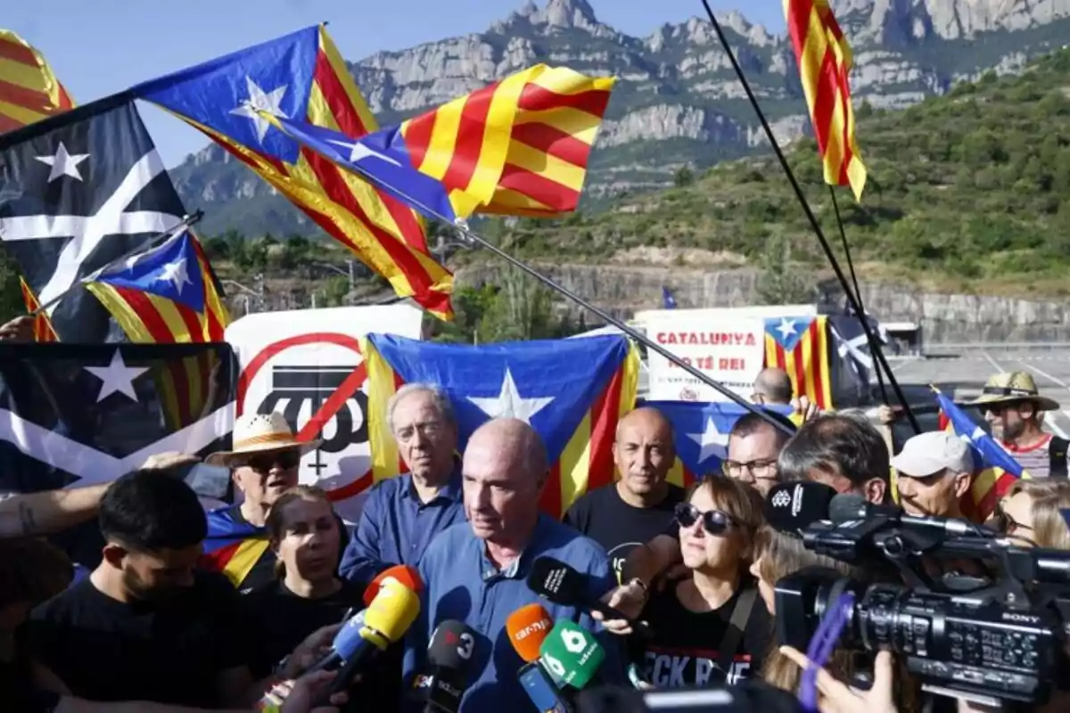 Un grupo de personas se manifiesta al aire libre con banderas esteladas y micrófonos de varios medios de comunicación, con montañas al fondo. Un grupo de personas se manifiesta al aire libre con banderas esteladas y micrófonos de varios medios de comunicación, con montañas al fondo.