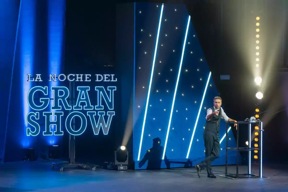Dani Martínez de pie en un escenario iluminado con luces azules y un letrero que dice La Noche del Gran Show Dani Martínez de pie en un escenario iluminado con luces azules y un letrero que dice La Noche del Gran Show