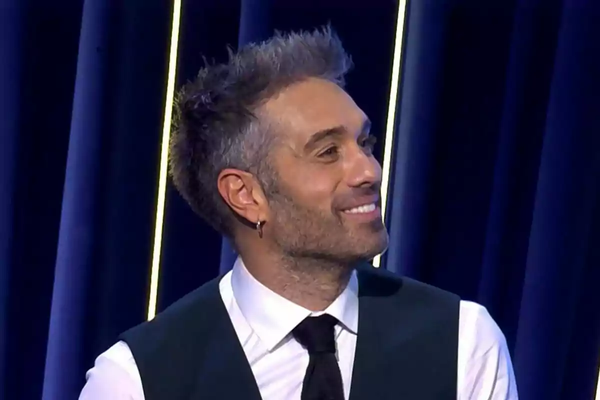 Dani Martínez de cabello corto y barba ligera sonriendo, vestido con camisa blanca, chaleco oscuro y corbata negra, frente a un fondo azul con líneas verticales iluminadas en El Gran Show.