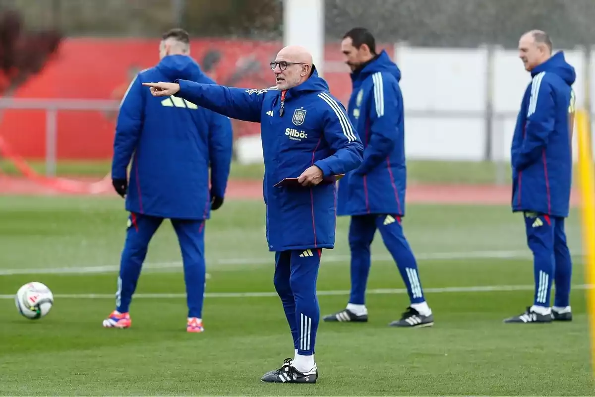 Entrenador de fútbol dando indicaciones en un campo de entrenamiento acompañado de otros miembros del cuerpo técnico, todos vestidos con ropa deportiva azul Entrenador de fútbol dando indicaciones en un campo de entrenamiento acompañado de otros miembros del cuerpo técnico, todos vestidos con ropa deportiva azul