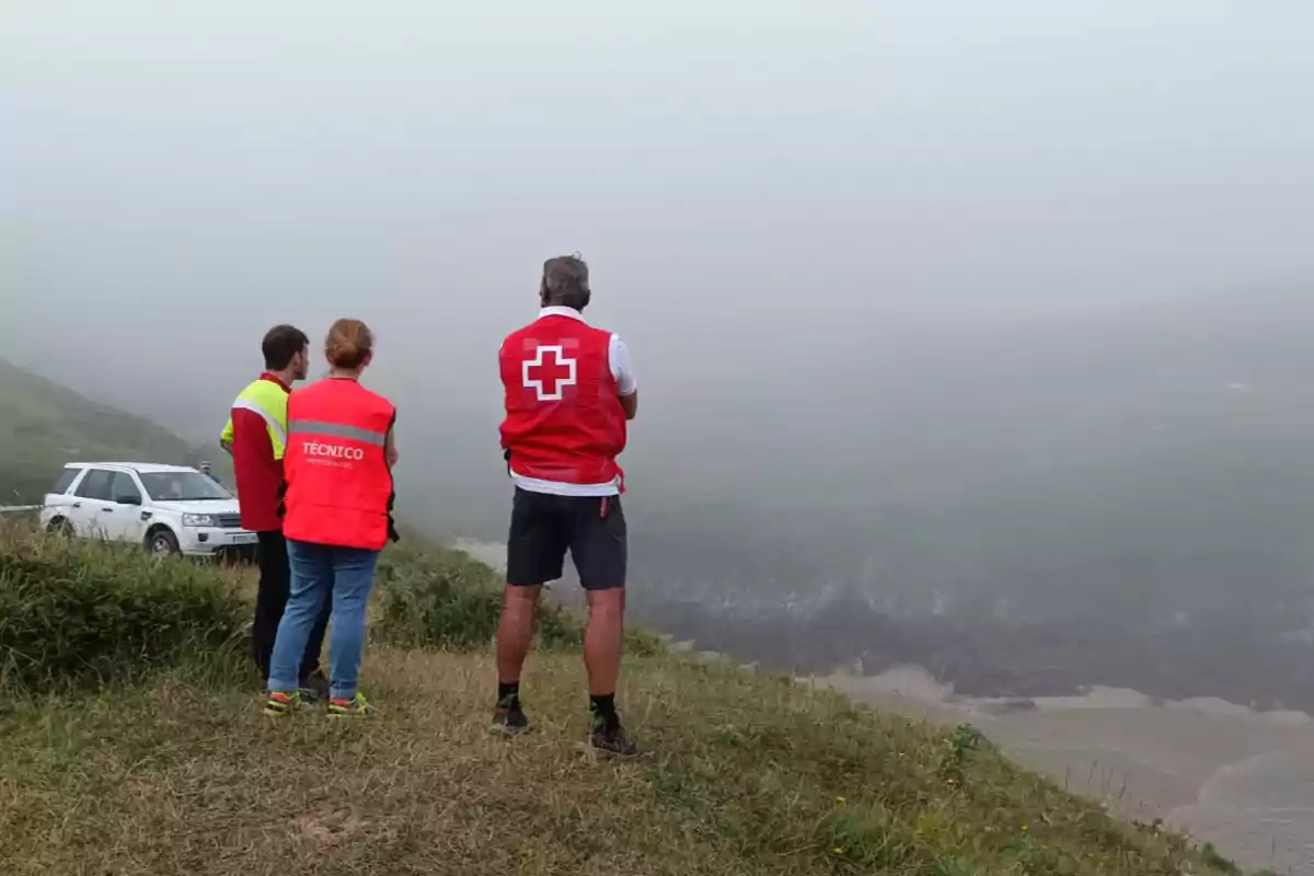 Tres personas con chalecos de emergencia observan un acantilado cubierto de niebla junto a un vehículo blanco estacionado en la hierba