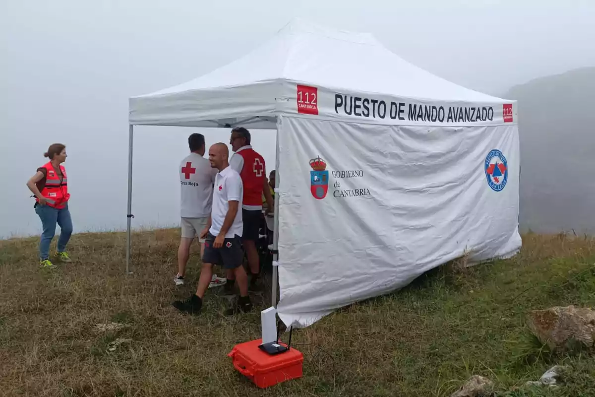 Varias personas con chalecos y camisetas de la Cruz Roja están reunidas junto a una carpa blanca de puesto de mando avanzado en una zona de campo con niebla. Varias personas con chalecos y camisetas de la Cruz Roja están reunidas junto a una carpa blanca de puesto de mando avanzado en una zona de campo con niebla.