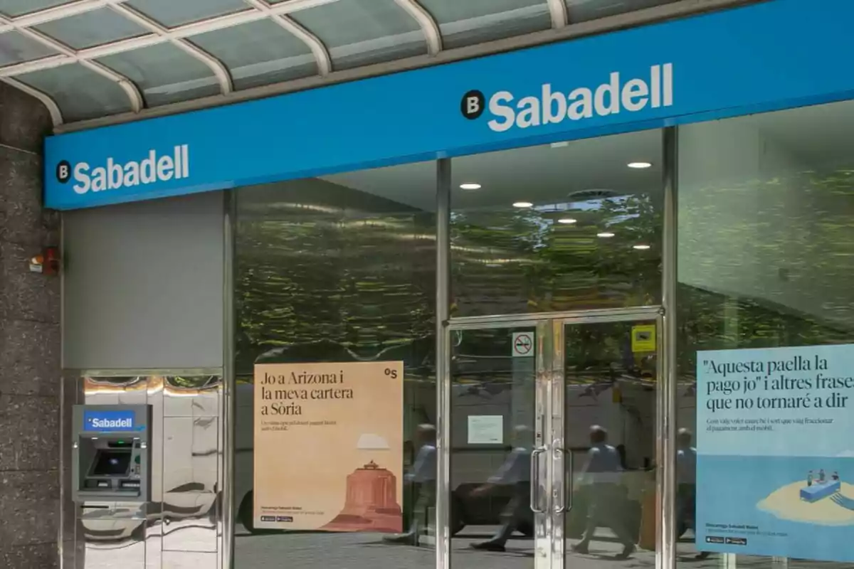 Fachada de una sucursal del banco Sabadell con un cajero automático y carteles publicitarios en las ventanas. Fachada de una sucursal del banco Sabadell con un cajero automático y carteles publicitarios en las ventanas.
