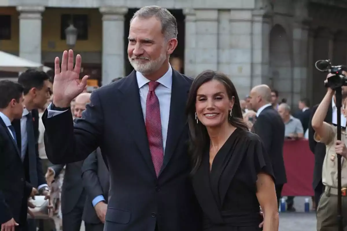 Felipe VI con traje oscuro y corbata roja saluda con la mano mientras la reina Letizia de vestido negro sonríe a su lado en un evento al aire libre rodeados de más personas Felipe VI con traje oscuro y corbata roja saluda con la mano mientras la reina Letizia de vestido negro sonríe a su lado en un evento al aire libre rodeados de más personas