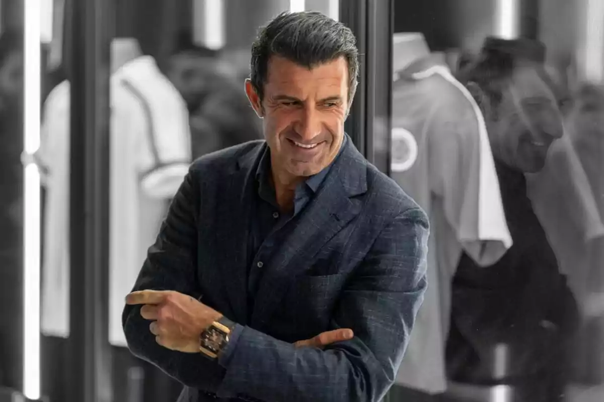 Hombre de cabello oscuro con saco azul sonriendo y señalando hacia un lado frente a un fondo con camisetas blancas y su reflejo en un vidrio Hombre de cabello oscuro con saco azul sonriendo y señalando hacia un lado frente a un fondo con camisetas blancas y su reflejo en un vidrio