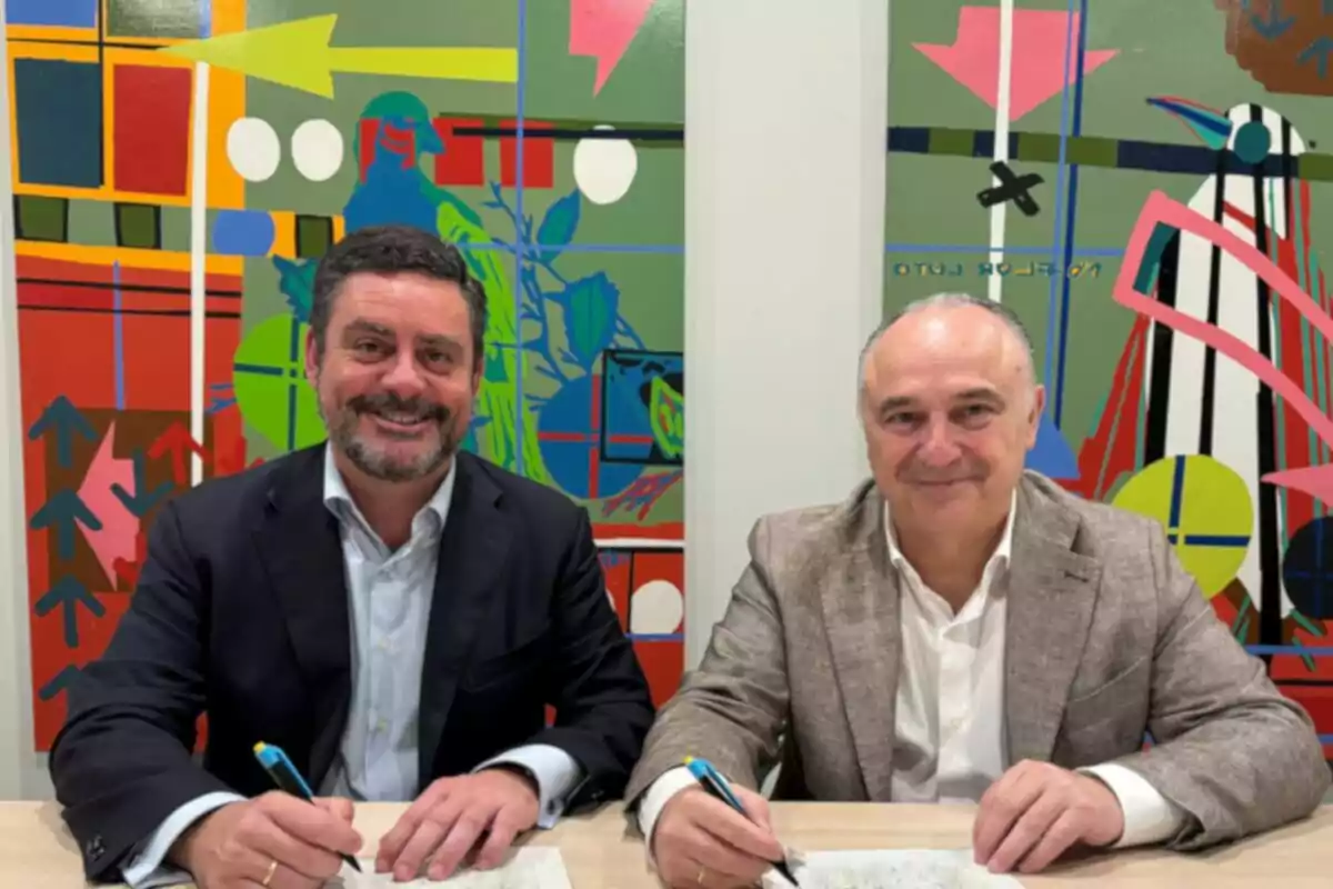 Dos hombres sonrientes sentados en una mesa firmando documentos con bolígrafos frente a un colorido mural abstracto Dos hombres sonrientes sentados en una mesa firmando documentos con bolígrafos frente a un colorido mural abstracto