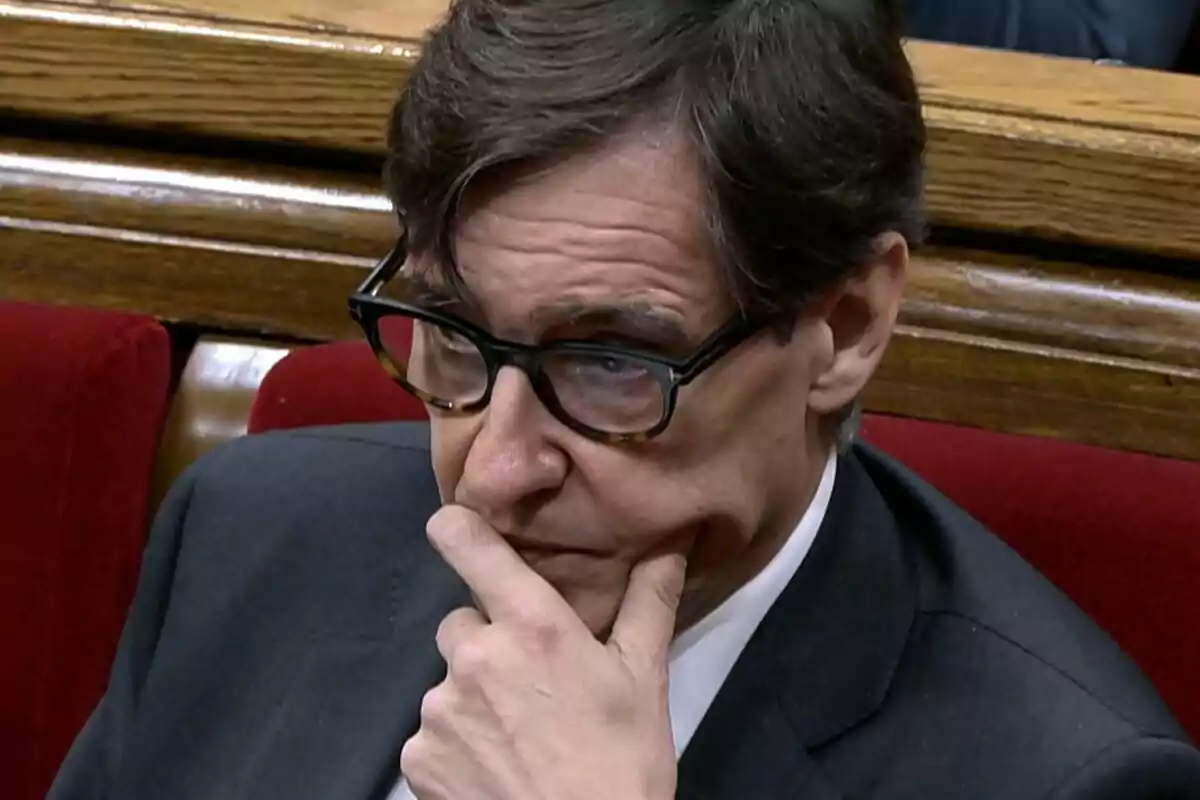 Hombre con gafas y traje oscuro sentado en un banco rojo con expresión pensativa y la mano en el rostro
