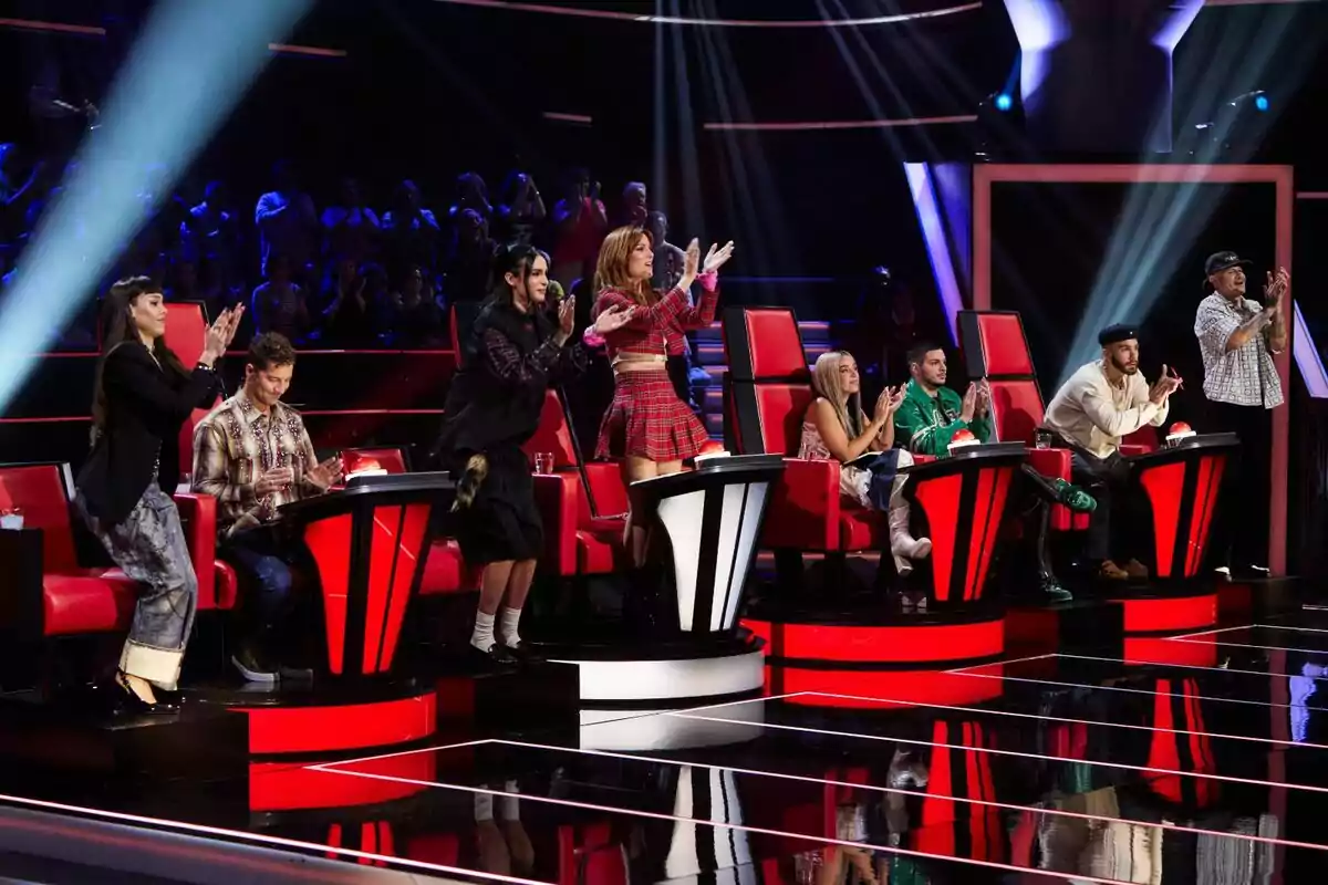 Varios jueces sentados y de pie aplauden en un set de televisión con sillas rojas y luces brillantes mientras el público observa al fondo de La Voz Kids Varios jueces sentados y de pie aplauden en un set de televisión con sillas rojas y luces brillantes mientras el público observa al fondo de La Voz Kids