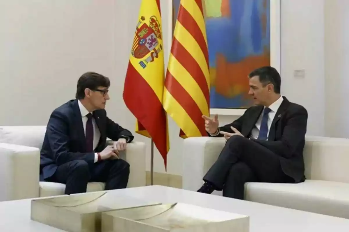 Dos hombres con traje conversan sentados en sillones blancos con las banderas de España y Cataluña detrás de ellos