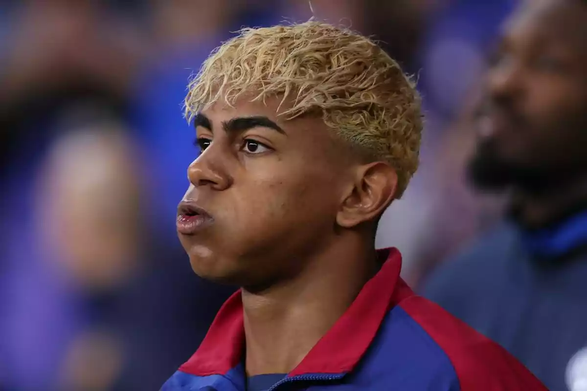 Joven con cabello rubio rizado y chaqueta deportiva azul y roja mirando hacia el frente con expresión seria Joven con cabello rubio rizado y chaqueta deportiva azul y roja mirando hacia el frente con expresión seria