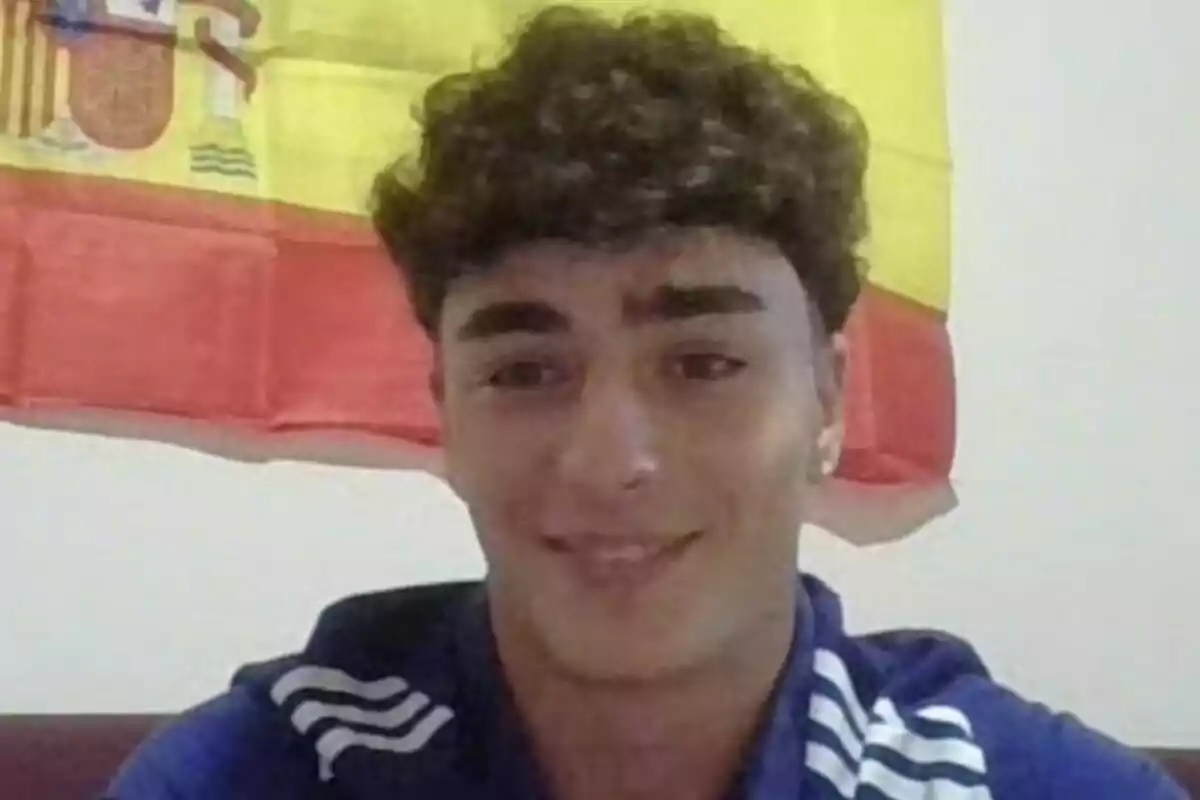 Joven con camiseta azul sonríe frente a una bandera de España en la pared.