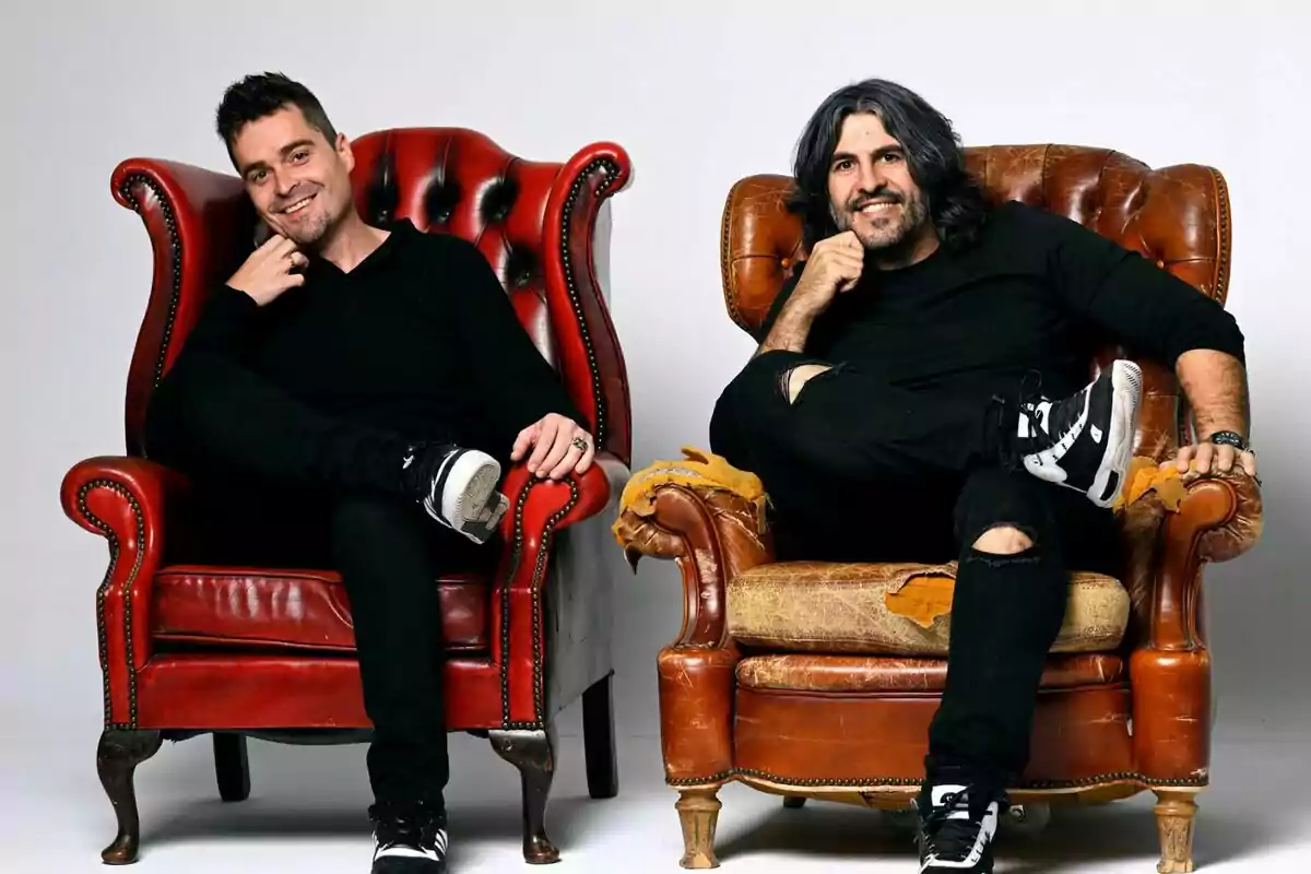 J.J. Vaquero y Álex Clavero sonrientes vestidos de negro están sentados en sillones antiguos, uno rojo y uno marrón, ambos con una pierna cruzada y la mano en la barbilla en La Garita J.J. Vaquero y Álex Clavero sonrientes vestidos de negro están sentados en sillones antiguos, uno rojo y uno marrón, ambos con una pierna cruzada y la mano en la barbilla en La Garita