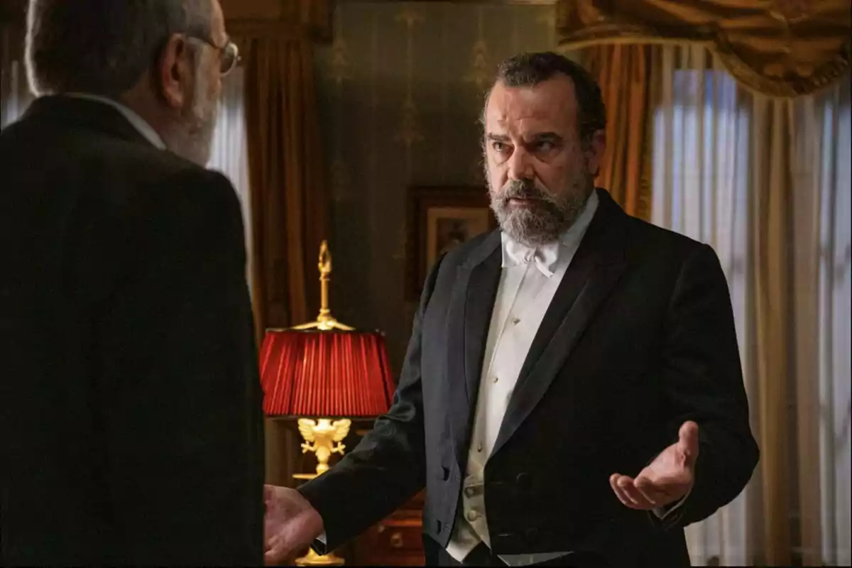 Alonso de barba con esmoquin blanco y negro gesticulando con las manos mientras habla con Rómulo en una habitación elegante con cortinas doradas y una lámpara de pantalla roja. Captura de 'La Promesa' Alonso de barba con esmoquin blanco y negro gesticulando con las manos mientras habla con Rómulo en una habitación elegante con cortinas doradas y una lámpara de pantalla roja. Captura de 'La Promesa'