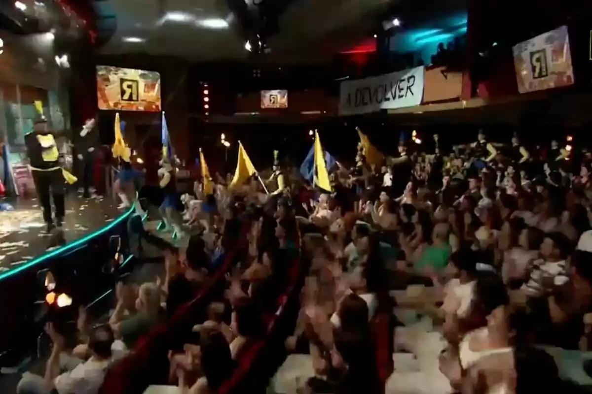Un teatro lleno de personas aplaudiendo mientras un grupo de animadores con banderas amarillas y azules se presenta en el escenario en 'La Revuelta'. Un teatro lleno de personas aplaudiendo mientras un grupo de animadores con banderas amarillas y azules se presenta en el escenario en 'La Revuelta'.