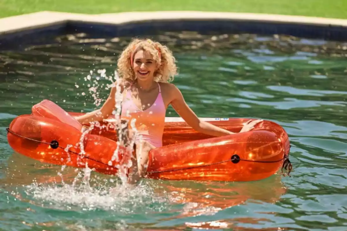 Mujer sonriente con traje de baño rosa sentada en un inflable naranja en una piscina mientras salpica agua Mujer sonriente con traje de baño rosa sentada en un inflable naranja en una piscina mientras salpica agua