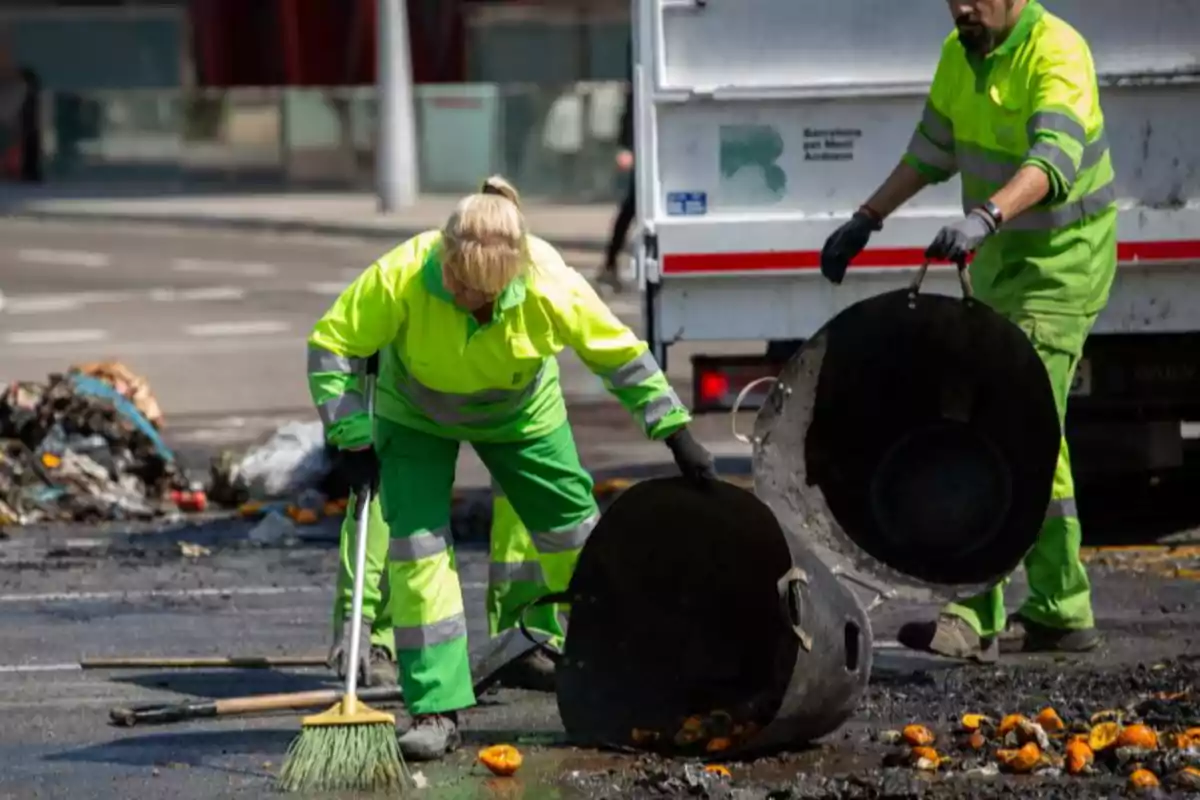 Dos trabajadores de limpieza con uniforme reflectante recogen basura y escombros en una calle usando cubos grandes y una escoba