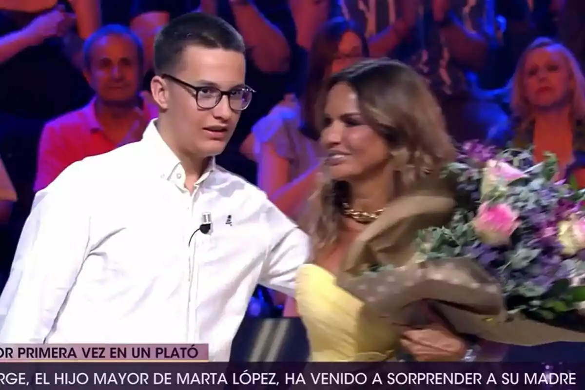 Marta López que sostiene un ramo de flores es abrazada por su hijo, un joven con gafas y camisa blanca, en un plató de De Viernes con público al fondo Marta López que sostiene un ramo de flores es abrazada por su hijo, un joven con gafas y camisa blanca, en un plató de De Viernes con público al fondo