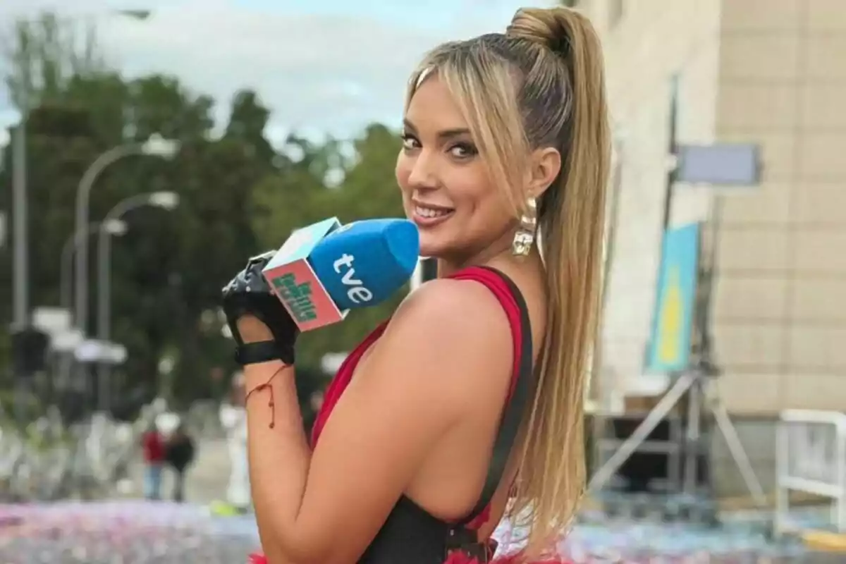 Marta Riesco rubia con coleta alta y vestido rojo sosteniendo un micrófono azul de tve mientras sonríe a la cámara en un evento al aire libre Marta Riesco rubia con coleta alta y vestido rojo sosteniendo un micrófono azul de tve mientras sonríe a la cámara en un evento al aire libre