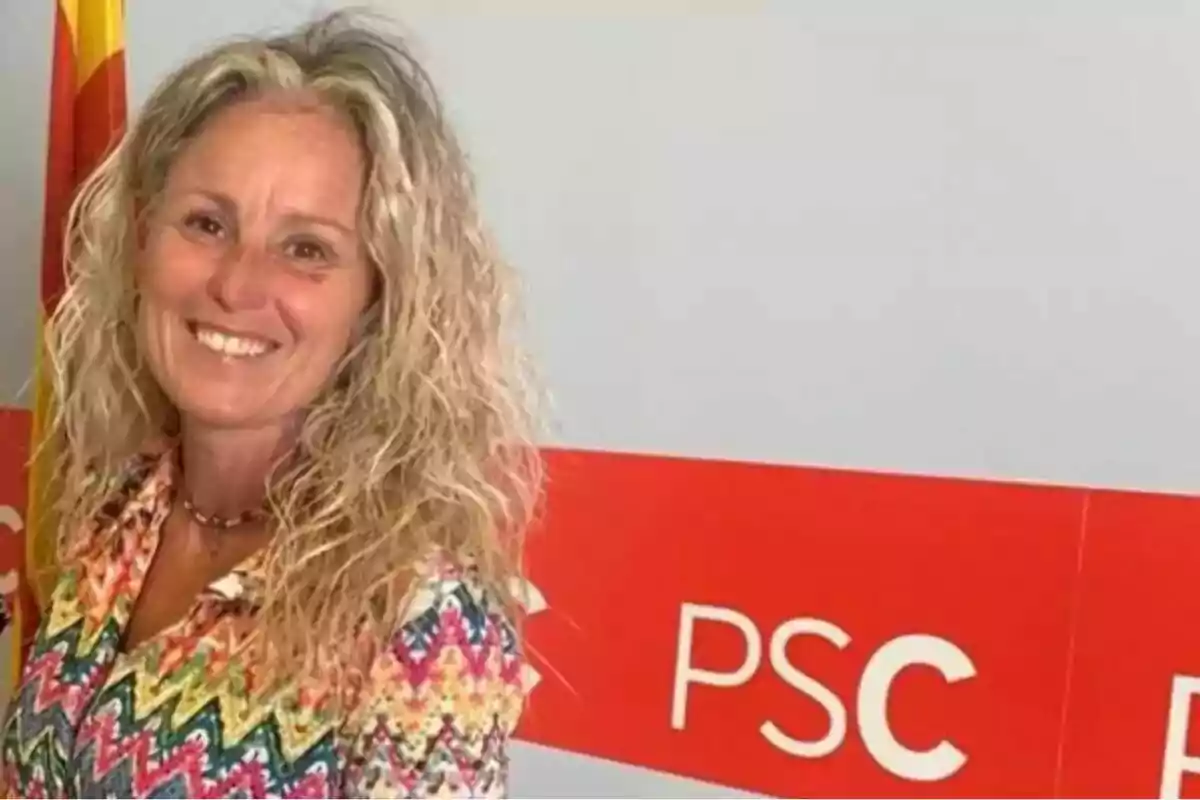 Mujer sonriente de cabello rubio rizado con camisa colorida posando frente a un fondo con el logo de PSC y una bandera. Mujer sonriente de cabello rubio rizado con camisa colorida posando frente a un fondo con el logo de PSC y una bandera.