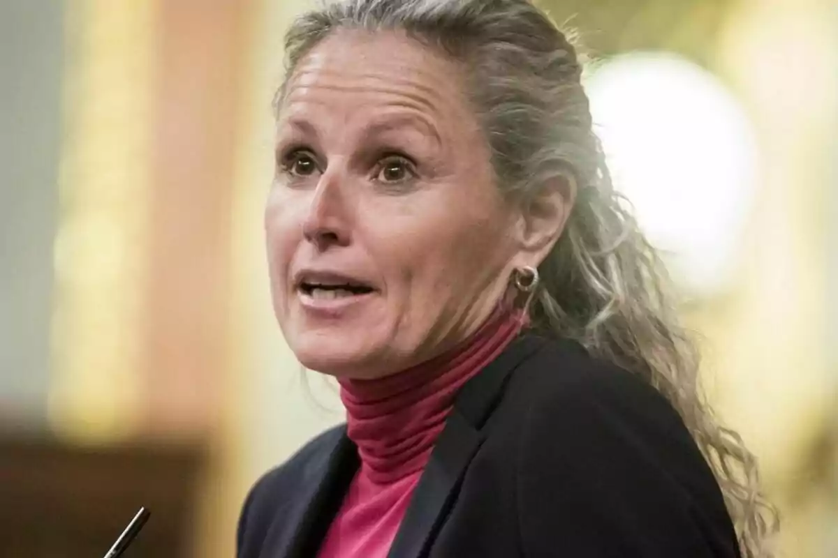 Mujer de cabello rubio y rizado recogido, usando un saco oscuro y suéter rosa, hablando en un entorno formal