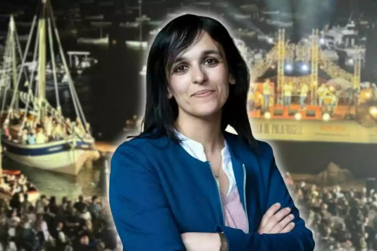 Mujer de cabello oscuro y chaqueta azul posando con los brazos cruzados frente a un fondo de un evento nocturno con barcos y un escenario iluminado