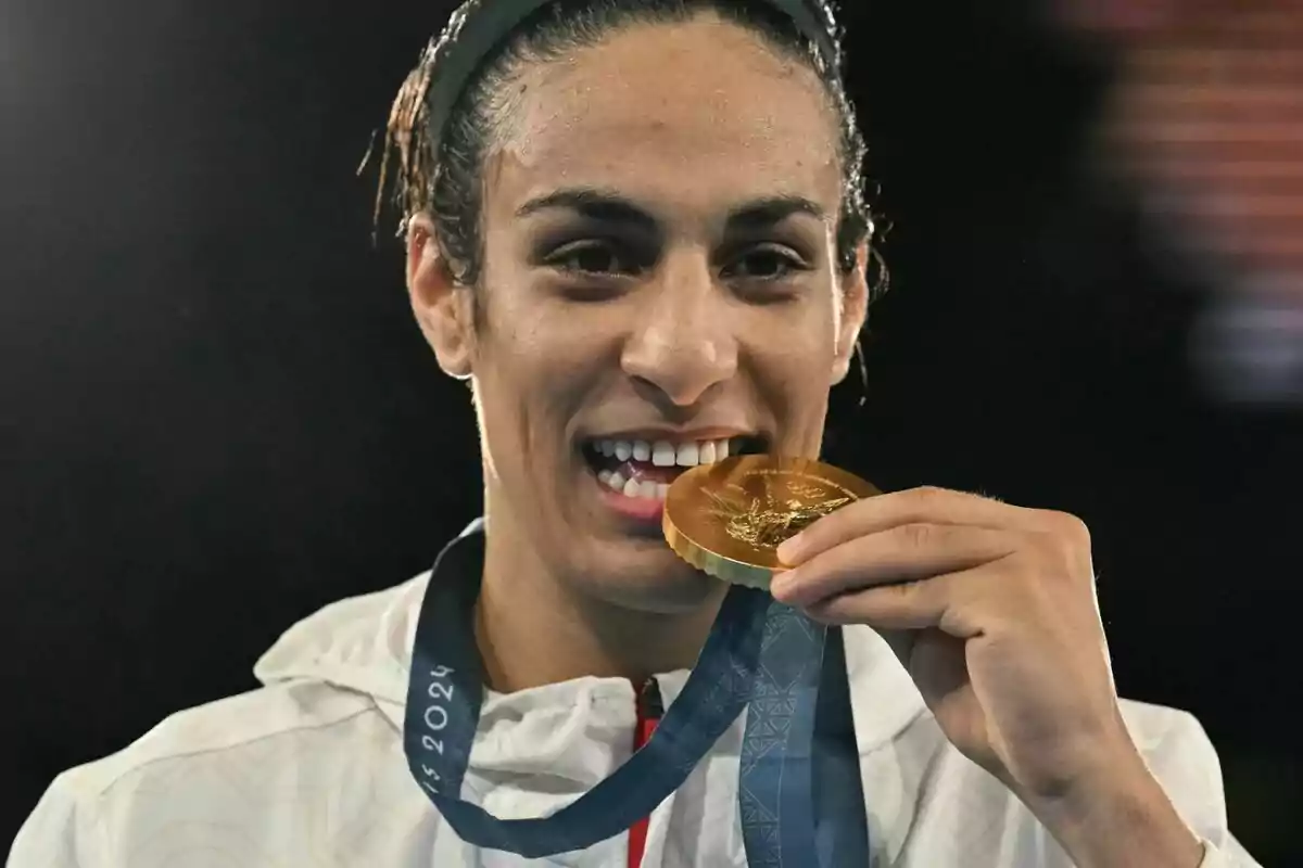 Persona sonriente muerde una medalla de oro mientras lleva una chaqueta blanca y una cinta azul