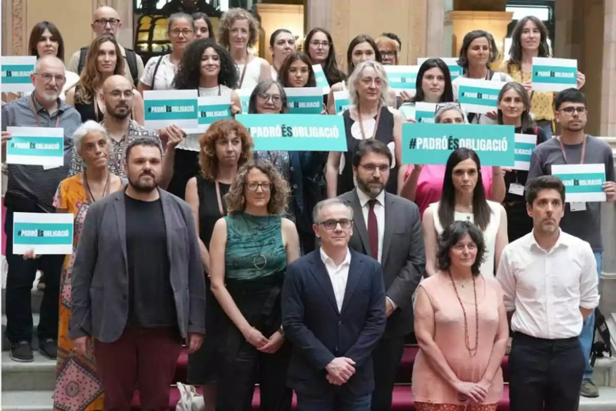 Un grupo de personas posando en una escalera, muchas de ellas sostienen carteles verdes con el mensaje PADRÓ ÉS OBLIGACIÓ Un grupo de personas posando en una escalera, muchas de ellas sostienen carteles verdes con el mensaje PADRÓ ÉS OBLIGACIÓ