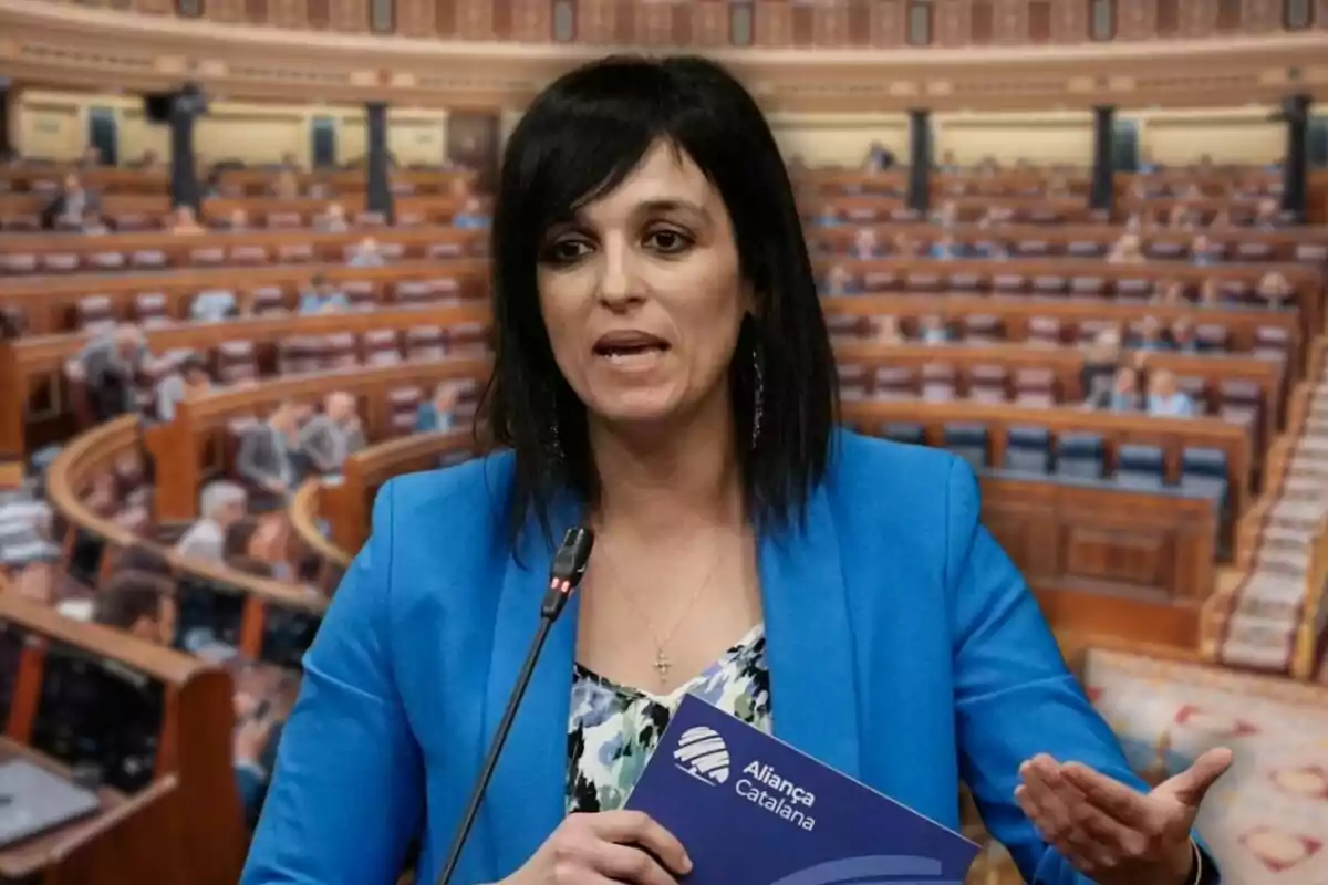 Mujer de cabello oscuro con chaqueta azul hablando en un parlamento mientras sostiene una carpeta de Aliança Catalana