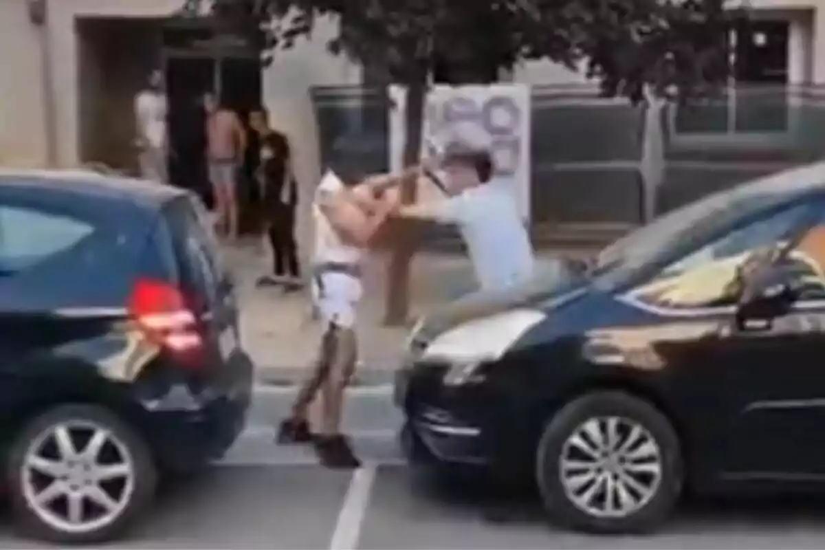 Dos personas discuten y se empujan entre dos autos estacionados en la calle mientras varias personas observan desde la acera.