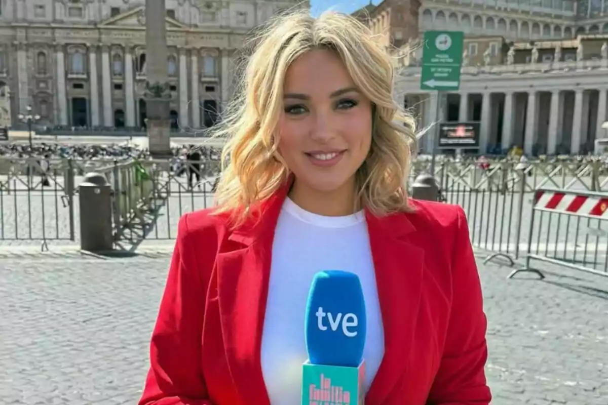 Marta Riesco rubia con chaqueta roja sostiene un micrófono azul de tve de 'La familia de la tele'