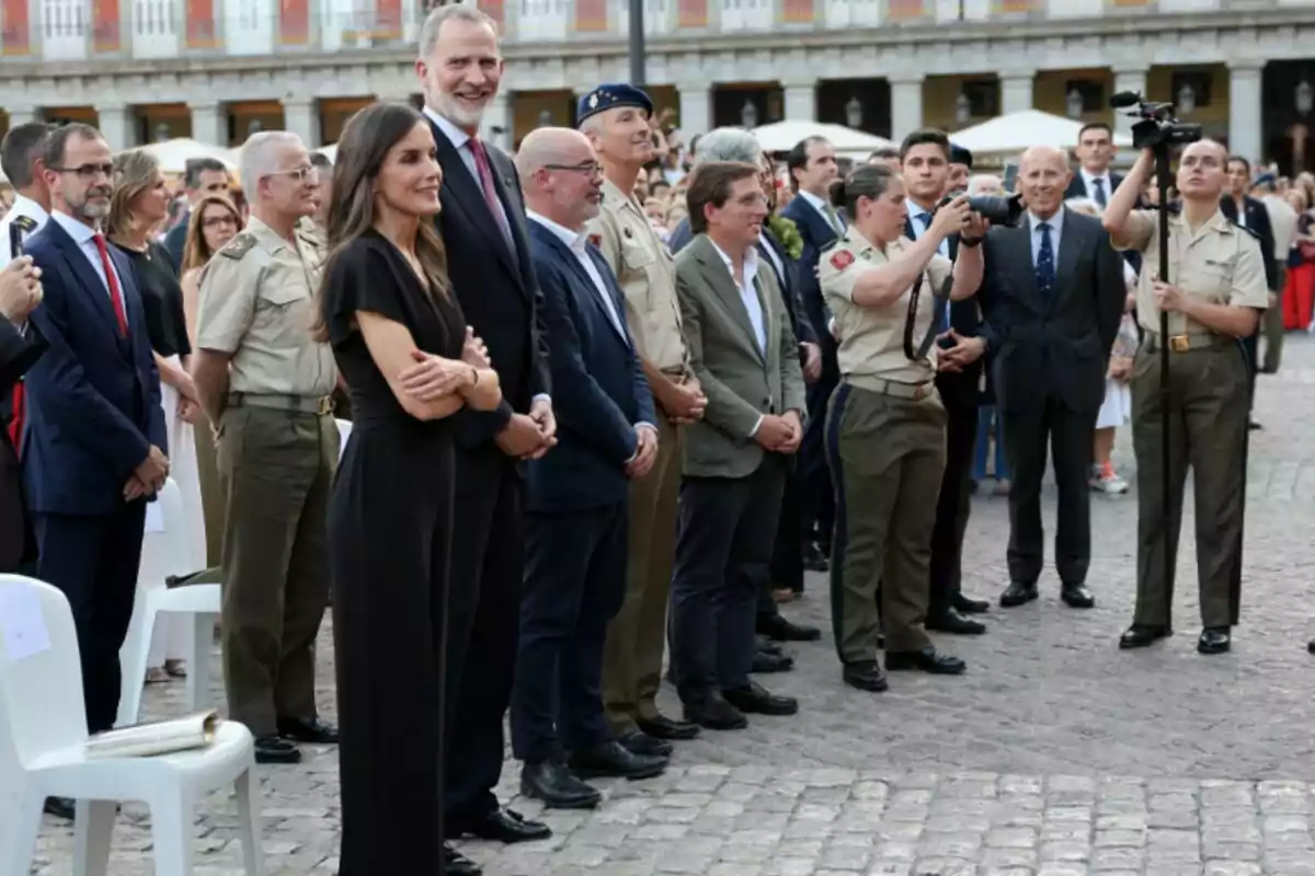 Un grupo de personas vestidas de manera formal y militar se encuentra de pie en una plaza al aire libre mientras observan y fotografían un evento Un grupo de personas vestidas de manera formal y militar se encuentra de pie en una plaza al aire libre mientras observan y fotografían un evento