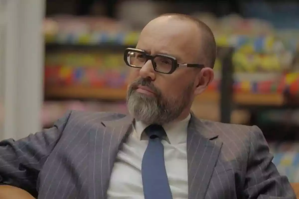 Risto Mejide con barba y gafas de pasta vistiendo un traje de rayas y corbata azul sentado en un ambiente interior desenfocado en 'Viajando con Chester'