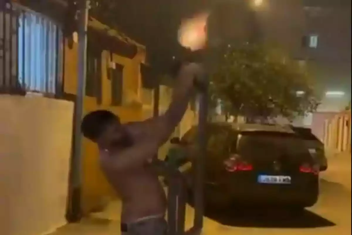 Hombre sin camisa disparando un arma larga hacia el cielo en una calle de noche