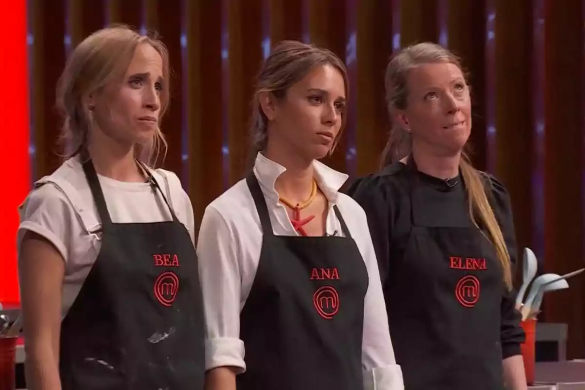 Tres mujeres con delantales negros de MasterChef y nombres bordados en rojo están de pie en una cocina con expresiones serias y concentradas