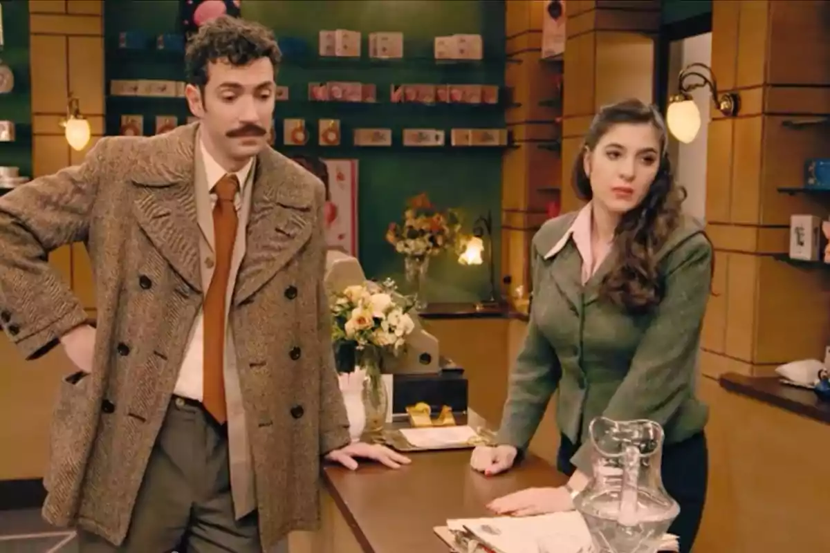 Un hombre con bigote y una mujer de cabello largo están apoyados en un mostrador dentro de una tienda decorada con flores y estanterías llenas de productos. Captura de 'Sueños de libertad'