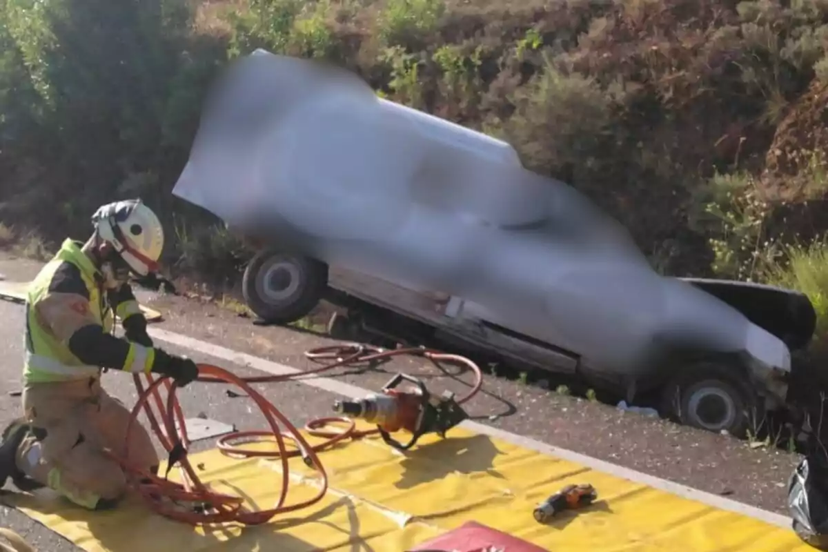 Bombero arrodillado en el suelo manipulando equipo de rescate junto a un vehículo accidentado fuera de la carretera en la A-1503, en Zaragoza.
